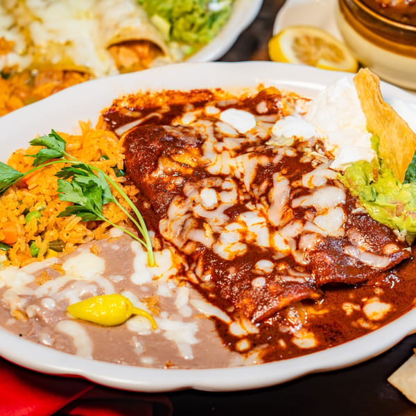 Plate of cheesy enchiladas with red sauce, rice, refried beans, guacamole, and sour cream.