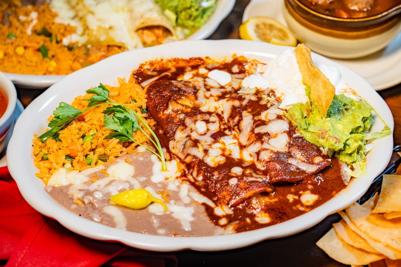 Cheesy enchiladas with red sauce, rice, refried beans, guacamole, sour cream, and a tortilla chip.