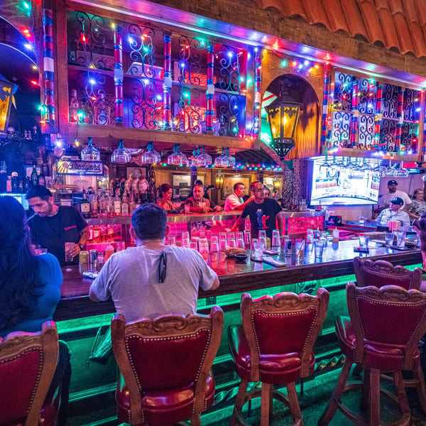 Patrons and bartenders at a vibrant, colorful bar with festive string lights.
