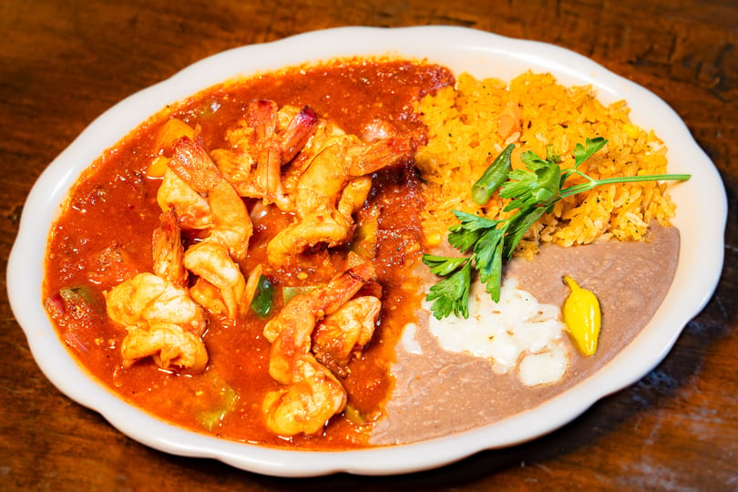 Spicy shrimp in red sauce, yellow rice, refried beans, and a yellow pepper on a white plate.