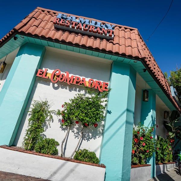 El Compadre Restaurant exterior with red tile roof, light blue walls, and vibrant plants under a clear sky.