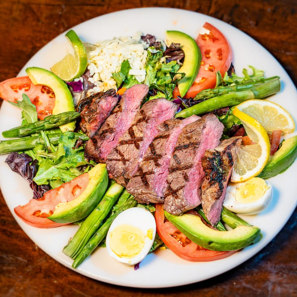 Vibrant grilled steak salad with avocado, asparagus, tomatoes, and egg on a white plate.