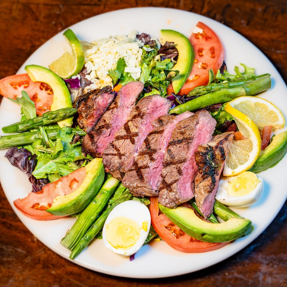 Vibrant grilled steak salad with avocado, asparagus, tomatoes, and egg on a white plate.
