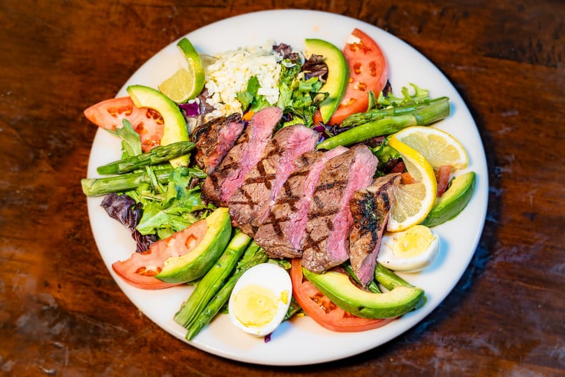 Vibrant grilled steak salad with avocado, tomatoes, asparagus, and egg on a white plate.