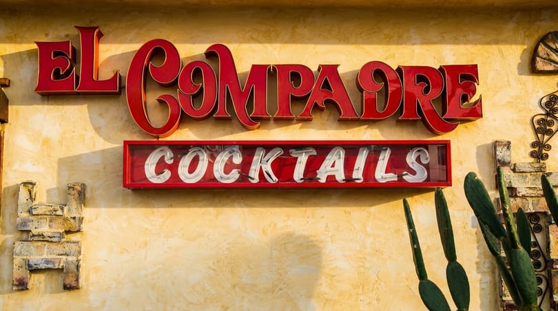 Red El Compadre sign and a neon "Cocktails" sign on a stucco wall with cacti.