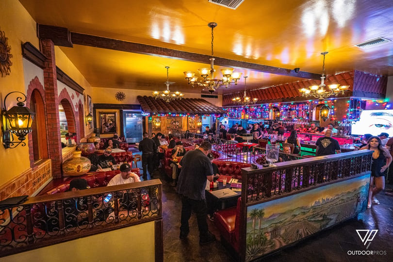 Lively restaurant interior with red booths, ornate chandeliers, and a bustling bar area with colorful lights.