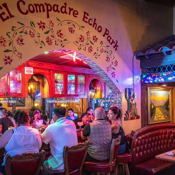 Lively bar area at El Compadre Echo Park with patrons, colorful lights, and painted archway.