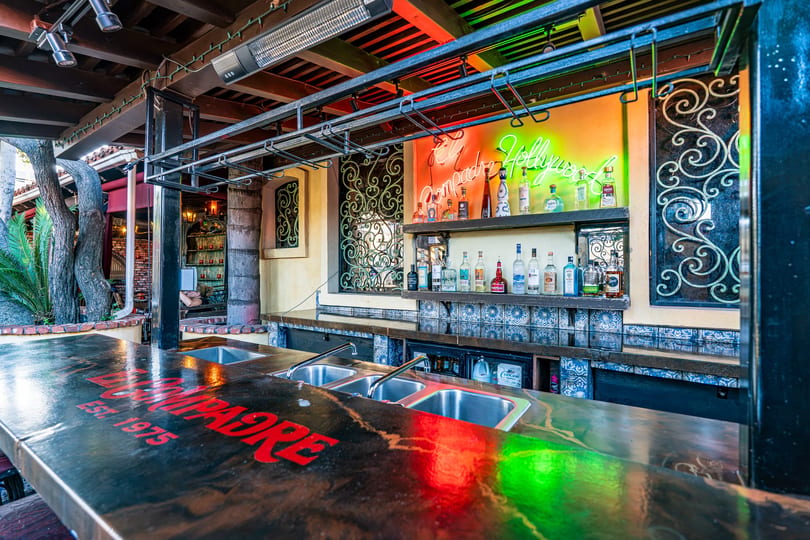 Vibrant outdoor bar with 'El Compadre Est. 1975' on the counter, neon sign, and liquor bottles.