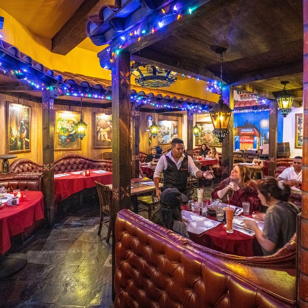 Lively restaurant interior with diners, a waiter, red tables, and colorful string lights.