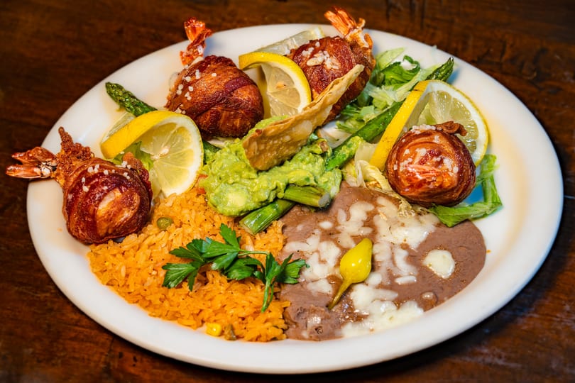 Bacon-wrapped shrimp with rice, refried beans, guacamole, asparagus, and lemon on a white plate.