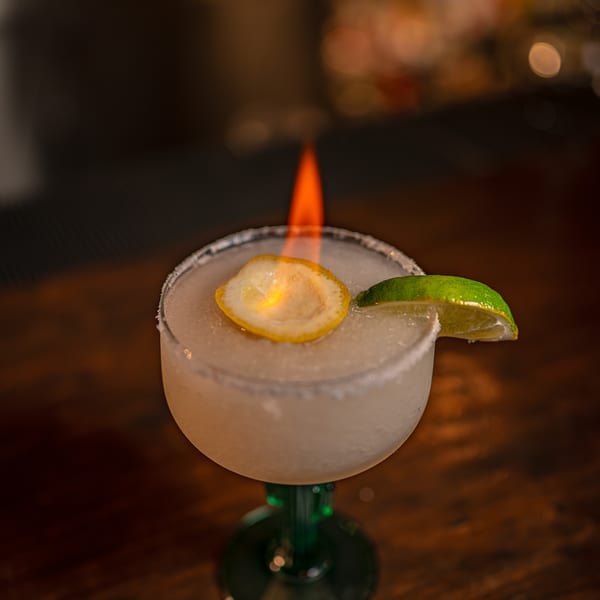 A flaming margarita with a lemon slice and lime wedge on a wooden bar.