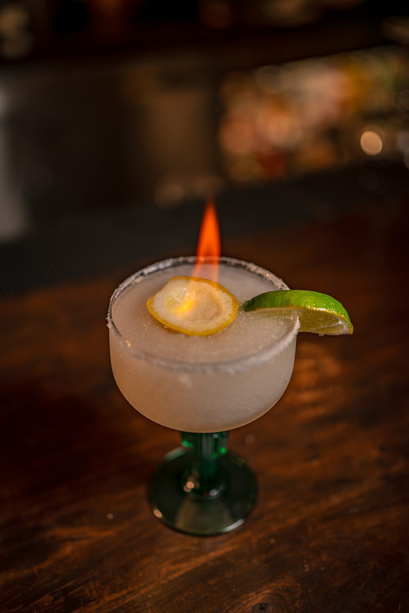 A flaming margarita with a lemon slice and lime wedge on a wooden bar.