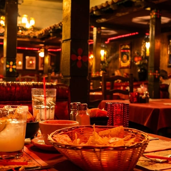 A dimly lit restaurant interior with red booths, tables set with drinks and chips, and patrons dining.