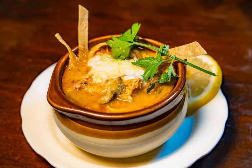 Hearty soup with melted cheese, tortilla strips, and parsley in a ceramic bowl with a lemon slice.
