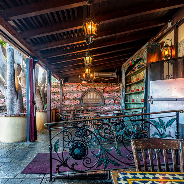 Outdoor restaurant patio with brick walls, large trees, ornate railings, and hanging lanterns.