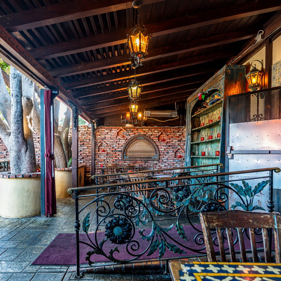 Outdoor restaurant patio with brick walls, large trees, ornate railings, and hanging lanterns.
