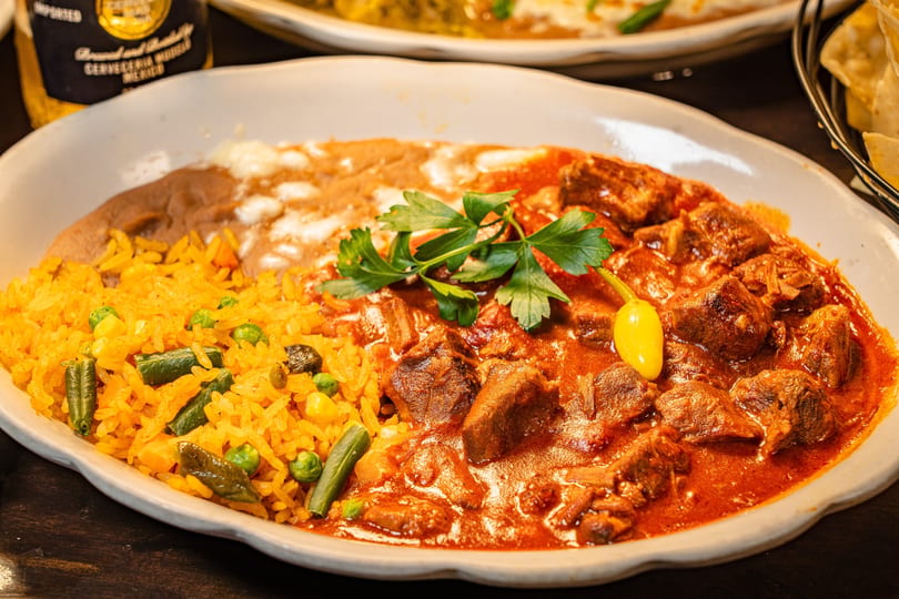 Hearty Mexican dish with stewed meat in red sauce, rice, refried beans, and a beer bottle.