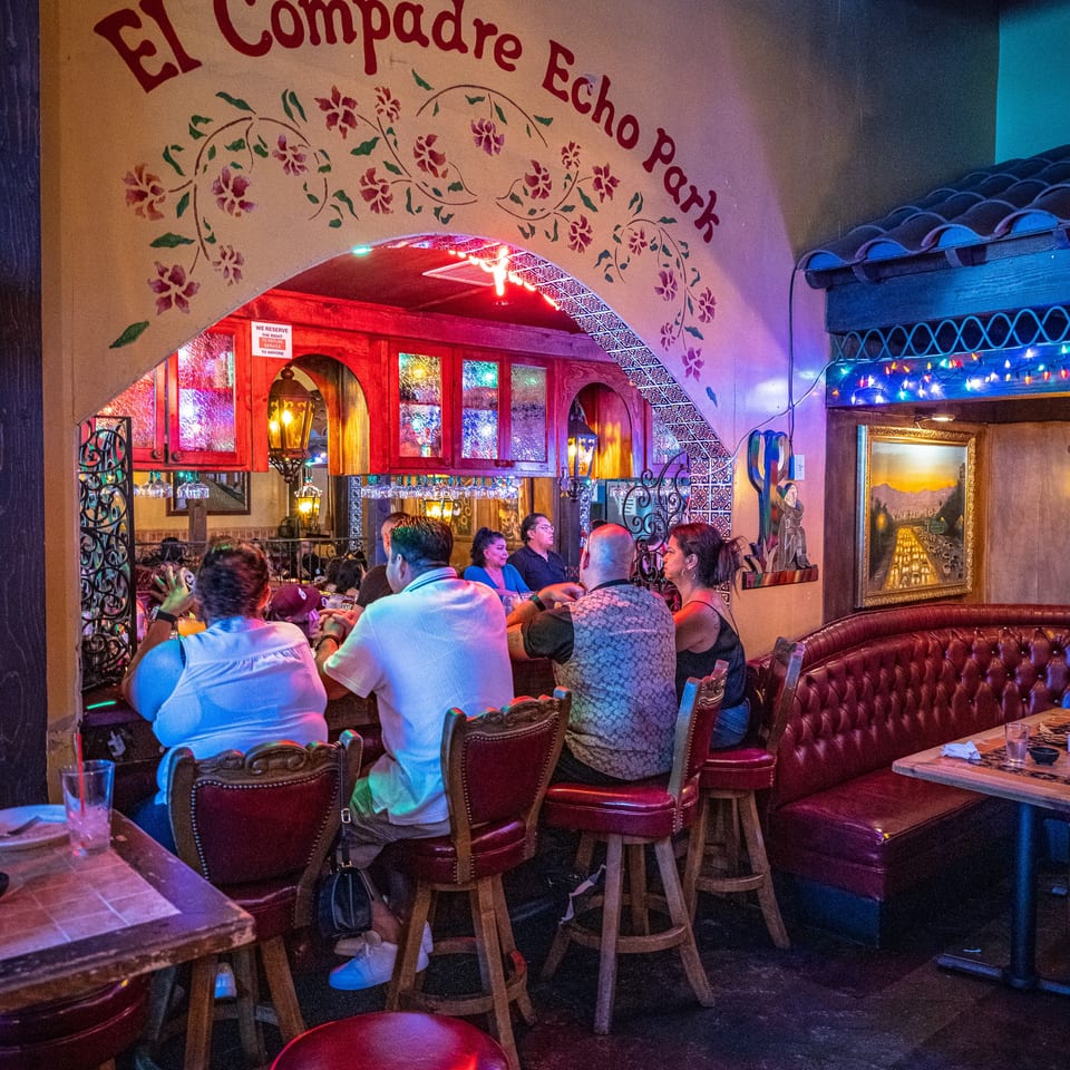 Patrons enjoy the vibrant, colorful interior of El Compadre Echo Park restaurant, featuring an arched entryway and bar seating.