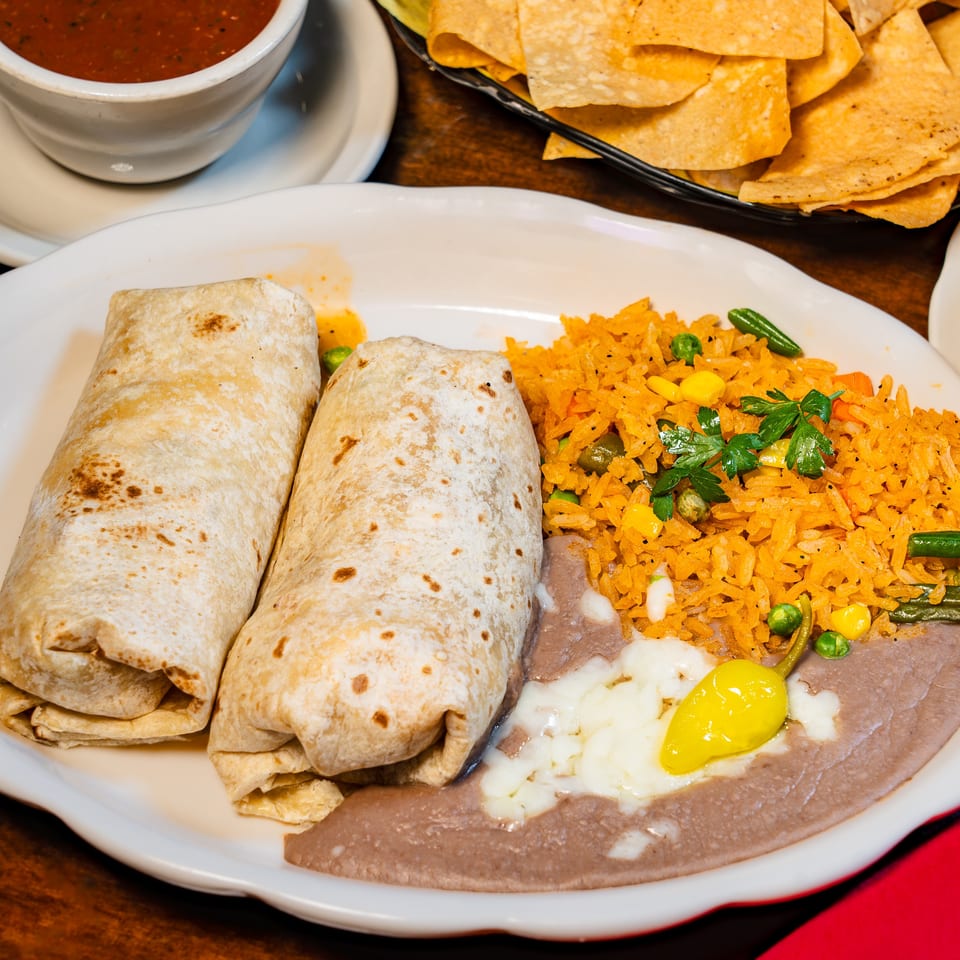 Two burritos, rice, and refried beans with cheese and a yellow pepper on a white plate.