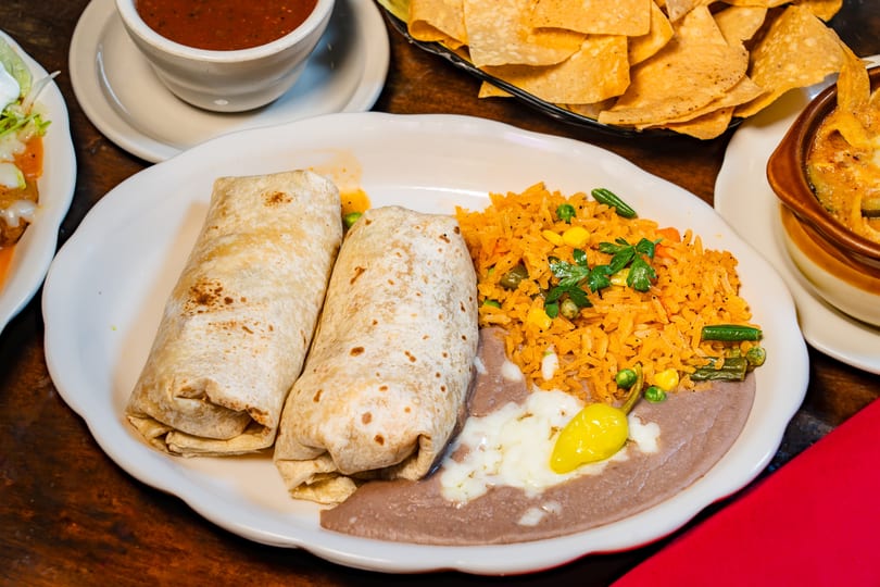 Two burritos with rice, refried beans, and a pepper on a white plate, with chips and salsa.