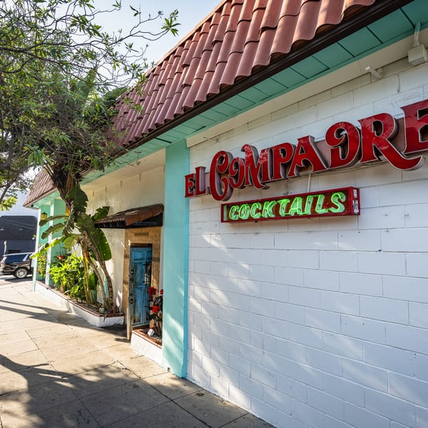 El Compadre restaurant exterior with a red tile roof, white walls, neon "Cocktails" sign, and sidewalk.