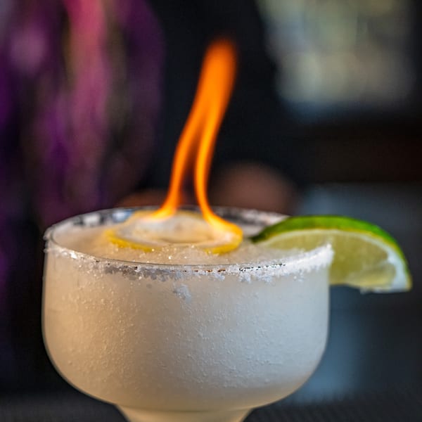 A flaming margarita with a salted rim, lemon, and lime garnish, served in a cactus-stemmed glass.