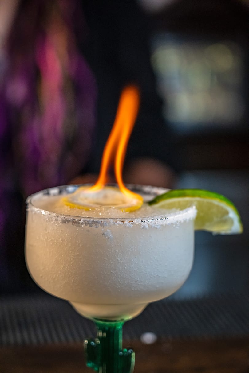 A flaming margarita with a salted rim, lemon, and lime garnish, served in a cactus-stemmed glass.