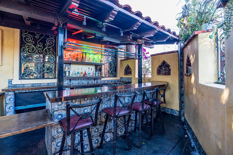 Vibrant outdoor bar with tiled counter, bar stools, and liquor bottles under a terracotta roof.