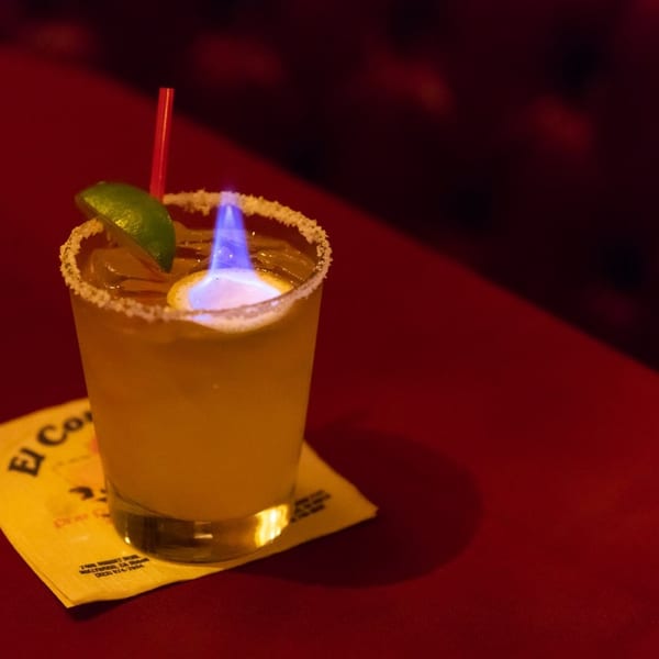 Flaming margarita with a lime wedge and red straw on a yellow napkin, on a red table.