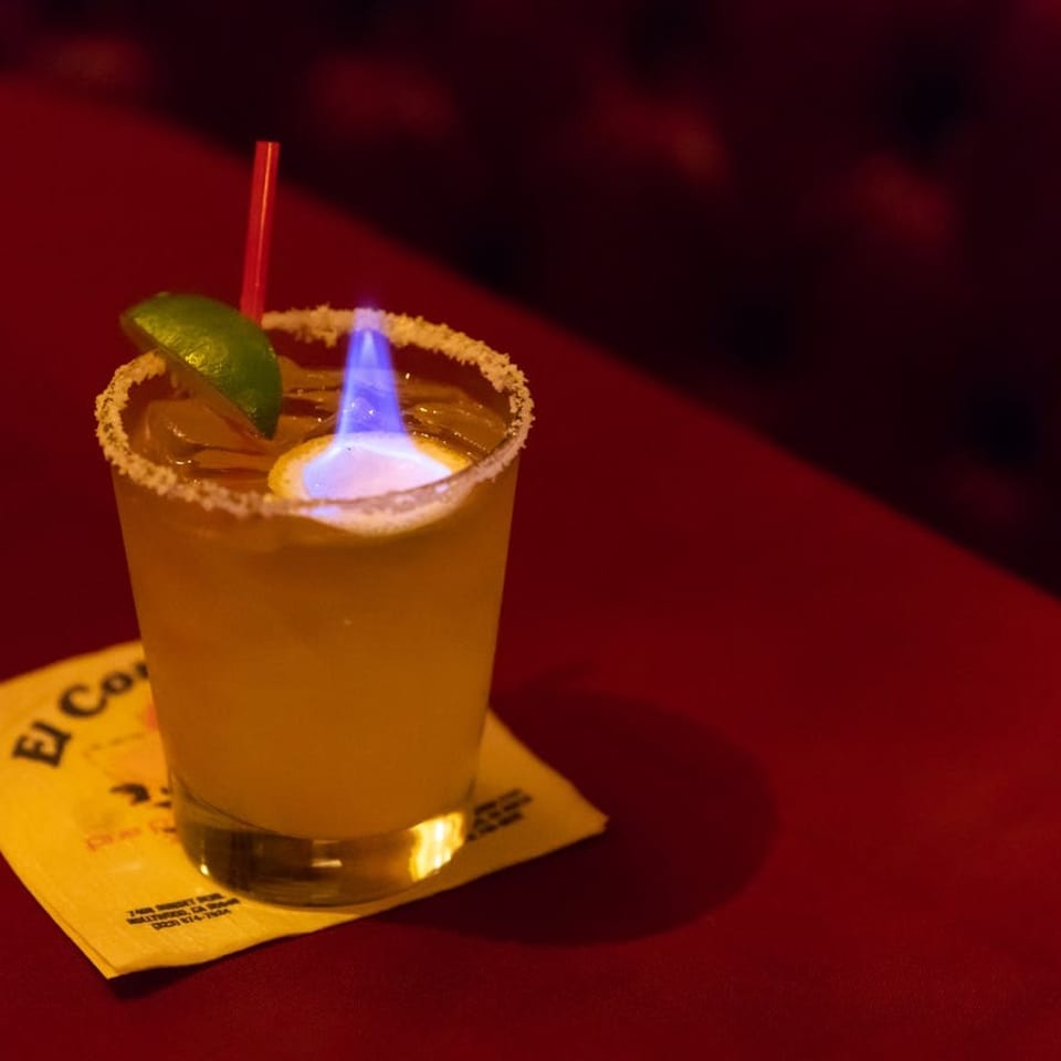 Flaming margarita with a lime wedge and red straw on a yellow napkin, on a red table.