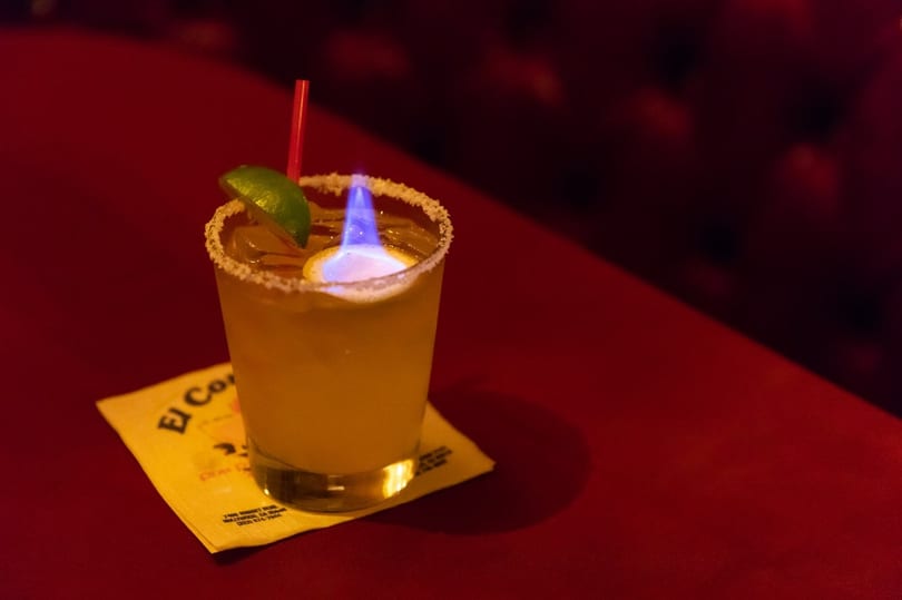 Flaming margarita with a lime wedge and red straw on a yellow napkin, on a red table.